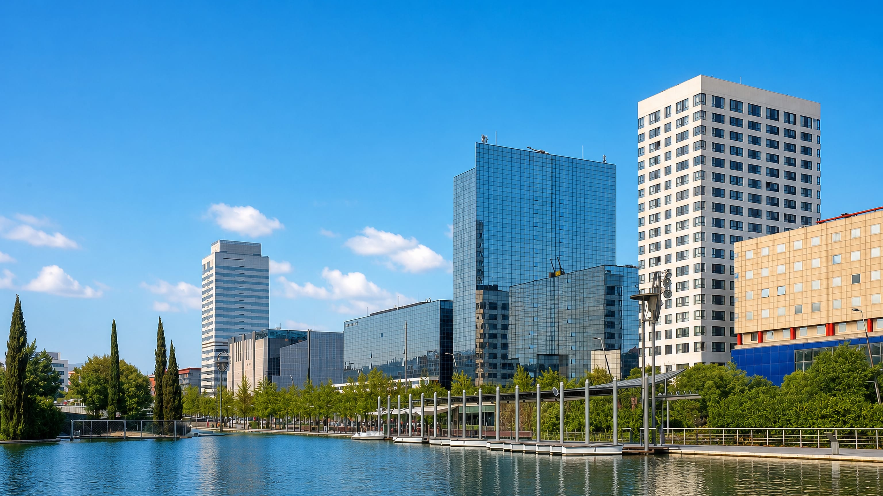 Glass office towers and a lakeside promenade in Sabadell, Catalonia, mirrored in still water under blue sky.