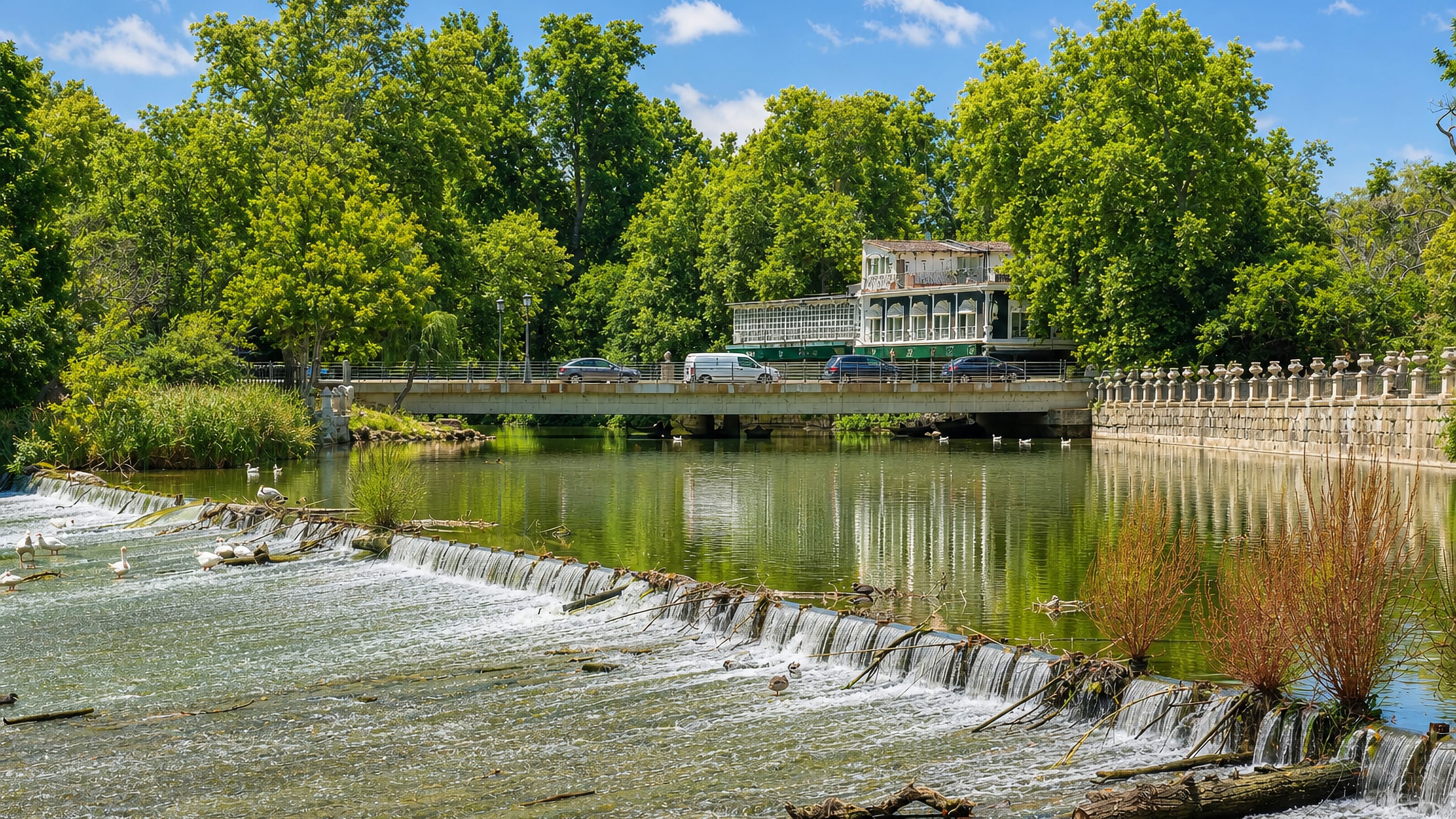 A weir on the Tagus at Aranjuez, with the balustraded garden embankment, a riverside pavilion and road bridge beyond