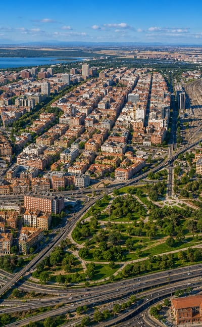 Aerial daytime panorama of Madrid's Arguelles district with a dense street grid, rail lines and a distant lake.