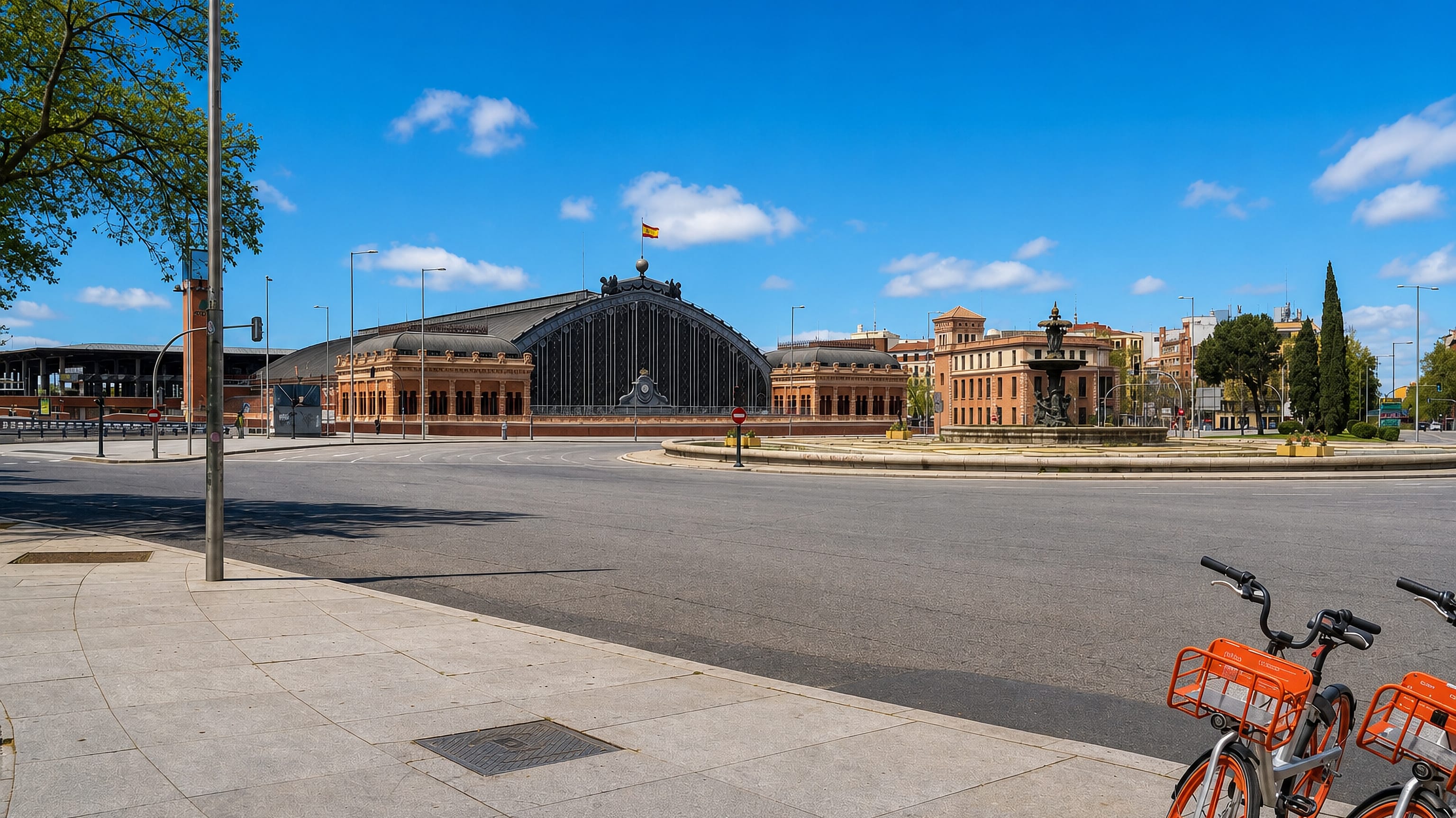 Atocha station's brick facade and arched iron train shed across a sunlit Madrid plaza with a stone fountain.