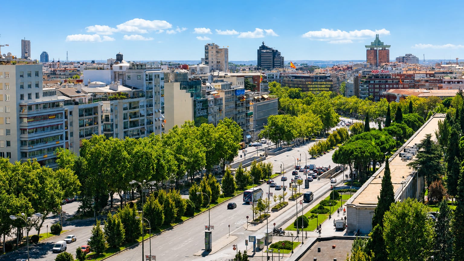 The wide tree-lined Paseo de la Castellana boulevard in Madrid, with a Spanish flag and mid-rise buildings under a bright sky.