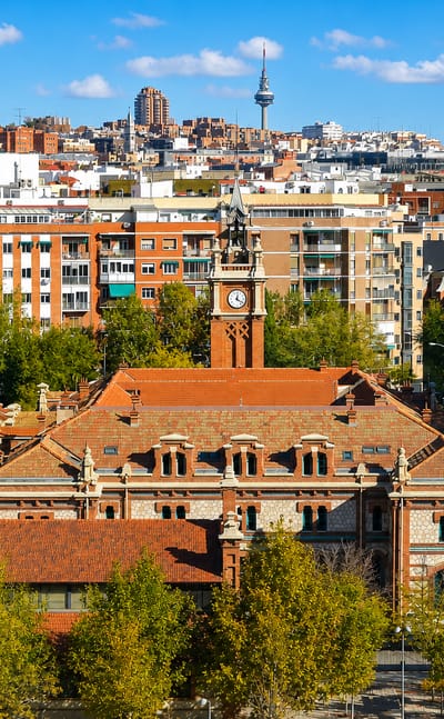 Red-brick historic complex with a clock tower and tree-lined boulevard in Chopera, Arganzuela, with Madrid's skyline and Torrespaña tower behind.