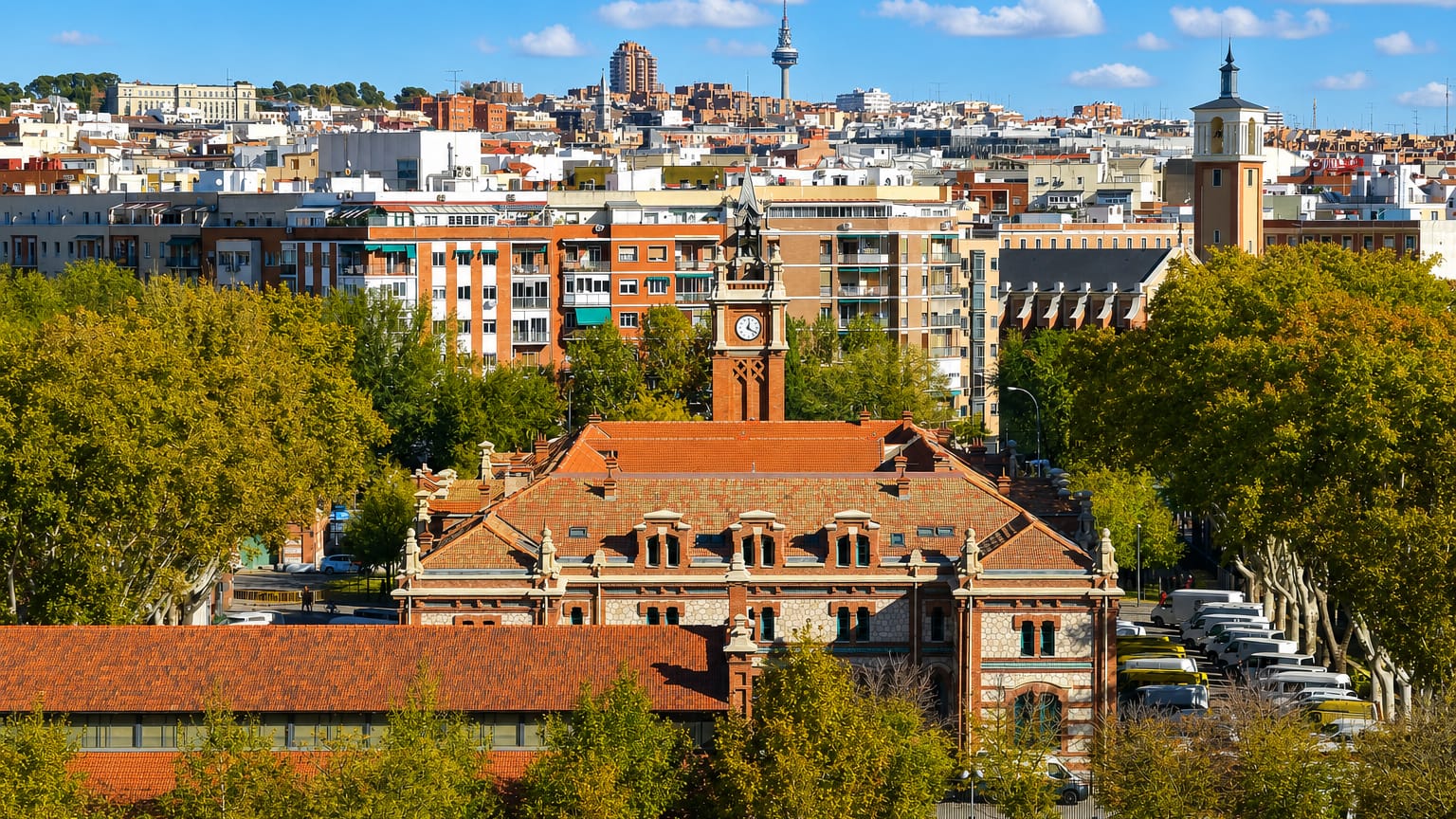 Red-brick historic complex with a clock tower and tree-lined boulevard in Chopera, Arganzuela, with Madrid's skyline and Torrespaña tower behind.