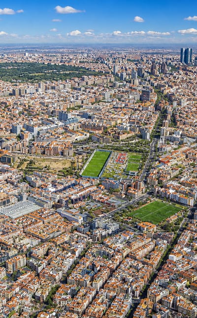 Aerial view across the dense street grid of Cuatro Caminos with Madrid's Cuatro Torres skyscrapers on the horizon.