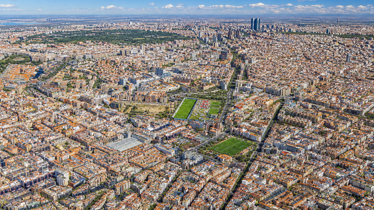 Aerial view across the dense street grid of Cuatro Caminos with Madrid's Cuatro Torres skyscrapers on the horizon.