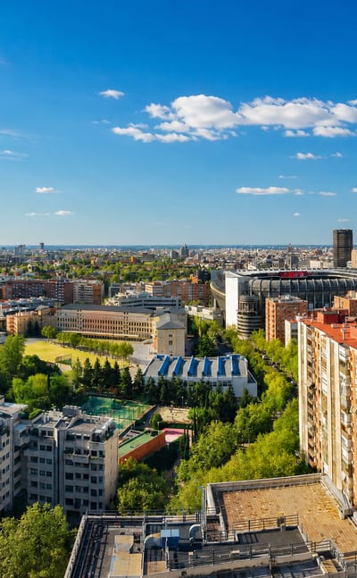 Aerial view over a dense Madrid residential district with green courtyards and the Cuatro Torres in the distance
