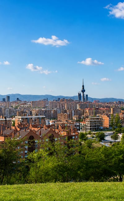 Madrid skyline seen from a green Vallecas hillside, with red-brick blocks and the Torrespaña tower against distant mountains.
