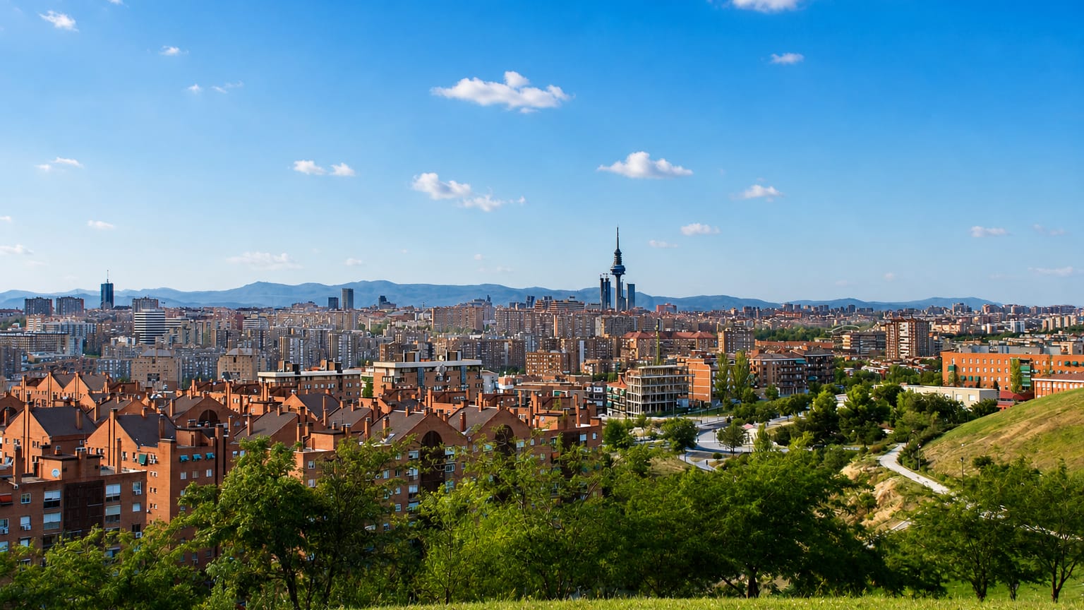 Madrid skyline seen from a green Vallecas hillside, with red-brick blocks and the Torrespaña tower against distant mountains.
