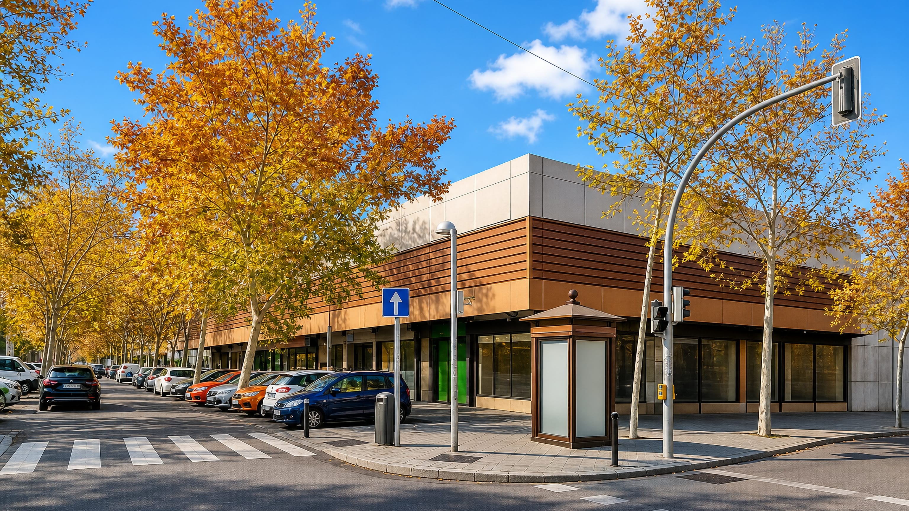 Sunlit street corner in Orcasitas, Madrid, with autumn plane trees, parked cars and a low brick-clad shopfront.