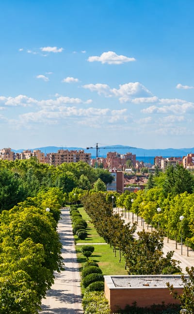 Green tree-lined park boulevard fronting mid-rise apartment blocks under a blue sky in Tetuán de las Victorias, Madrid