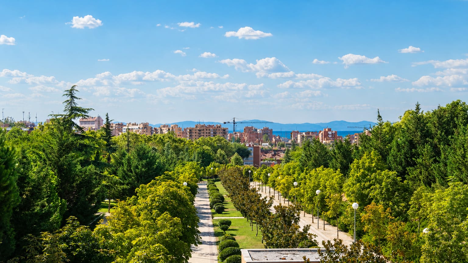 Green tree-lined park boulevard fronting mid-rise apartment blocks under a blue sky in Tetuán de las Victorias, Madrid
