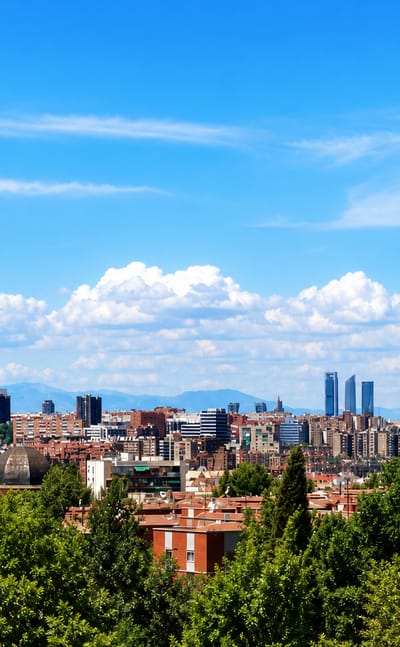 Panoramic Madrid skyline seen from a green hillside, with the Cuatro Torres and the Torrespana TV tower against a blue sky.