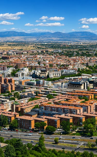 Aerial view of red-brick residential blocks and wide roads in the Valverde neighborhood of northern Madrid, with the Sierra de Guadarrama behind.