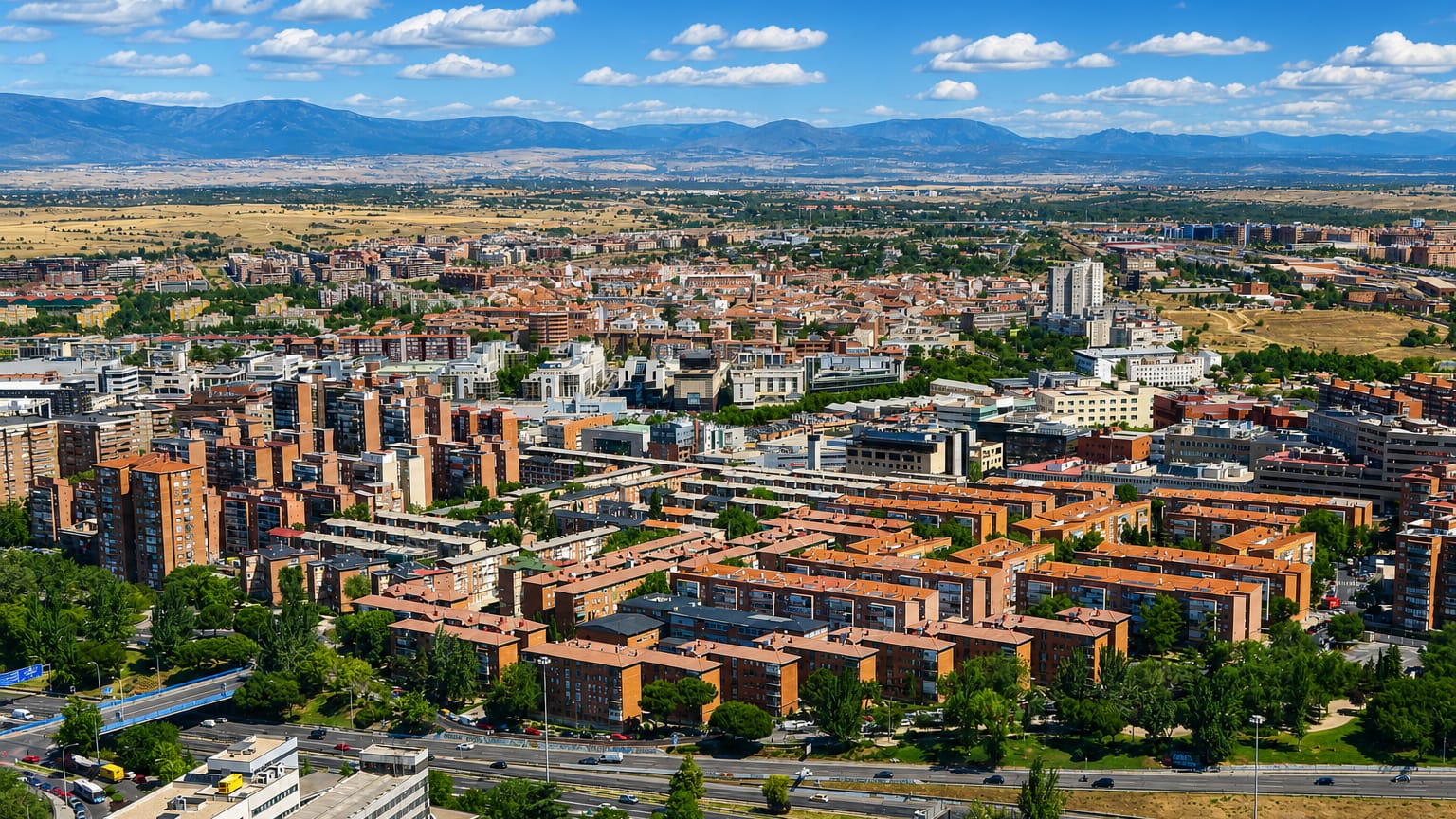 Aerial view of red-brick residential blocks and wide roads in the Valverde neighborhood of northern Madrid, with the Sierra de Guadarrama behind.