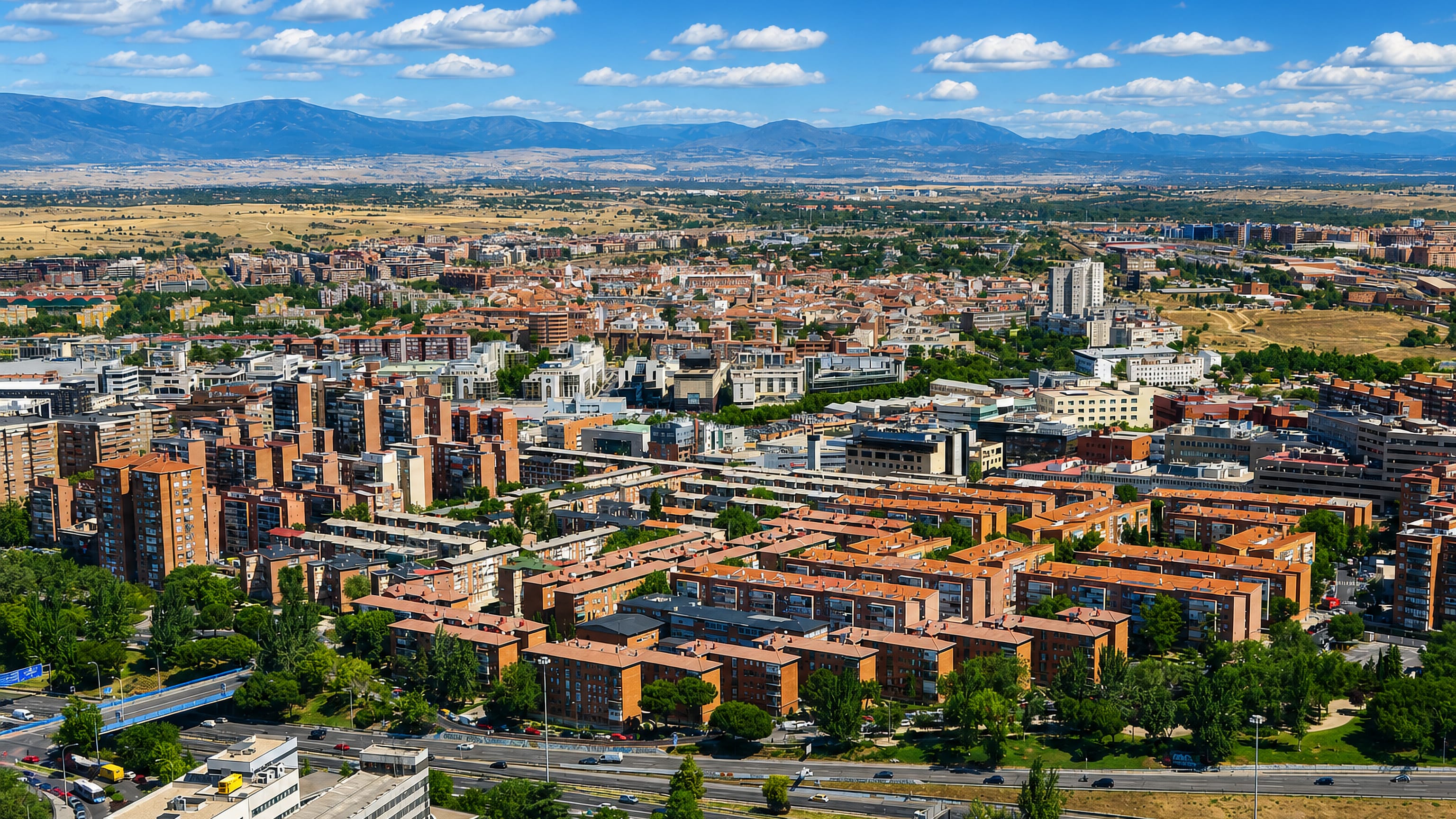 Aerial view of red-brick residential blocks and wide roads in the Valverde neighborhood of northern Madrid, with the Sierra de Guadarrama behind.