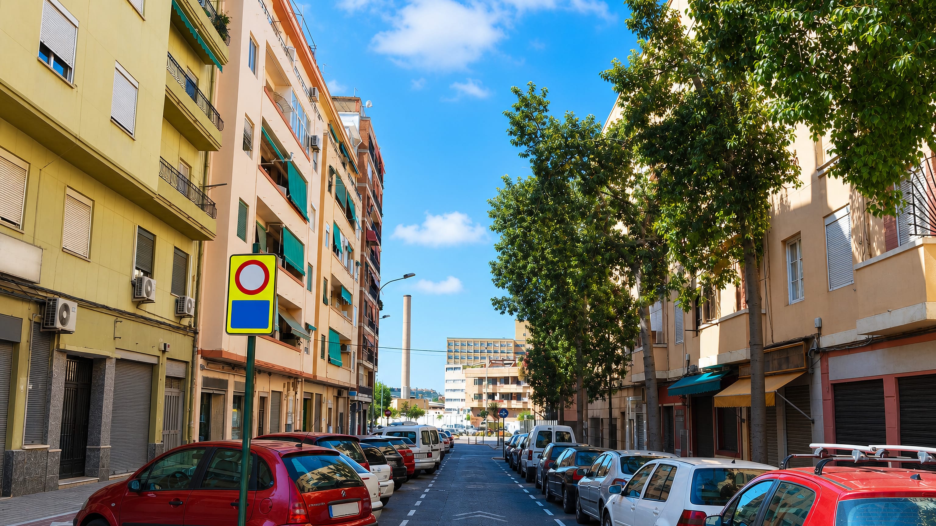 Residential street in El Calvari, Valencia, with parked cars, pastel apartment blocks and an old factory chimney beyond.