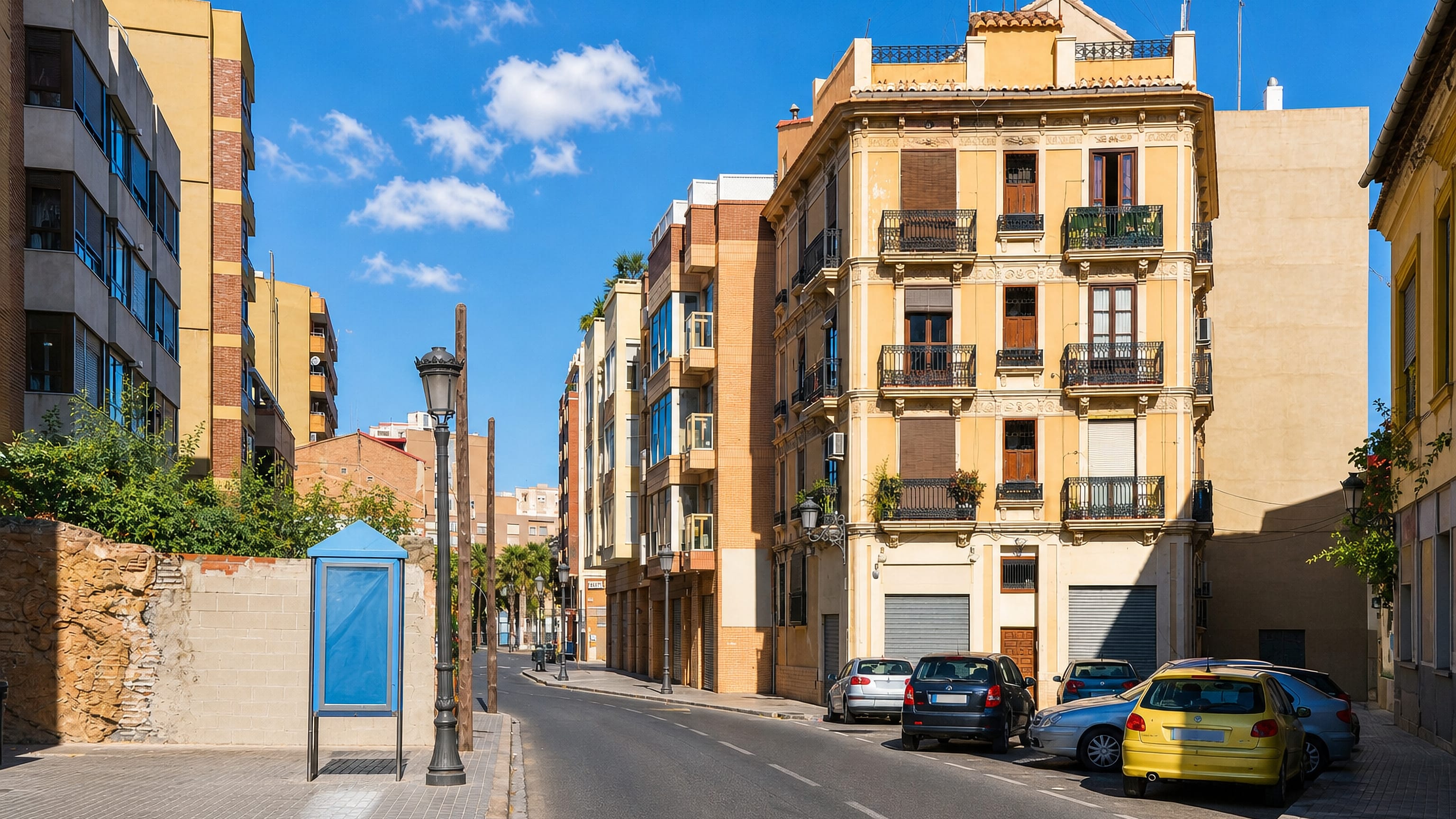 Sunlit street of yellow balconied apartment blocks with parked cars in Valencia's Morvedre district.