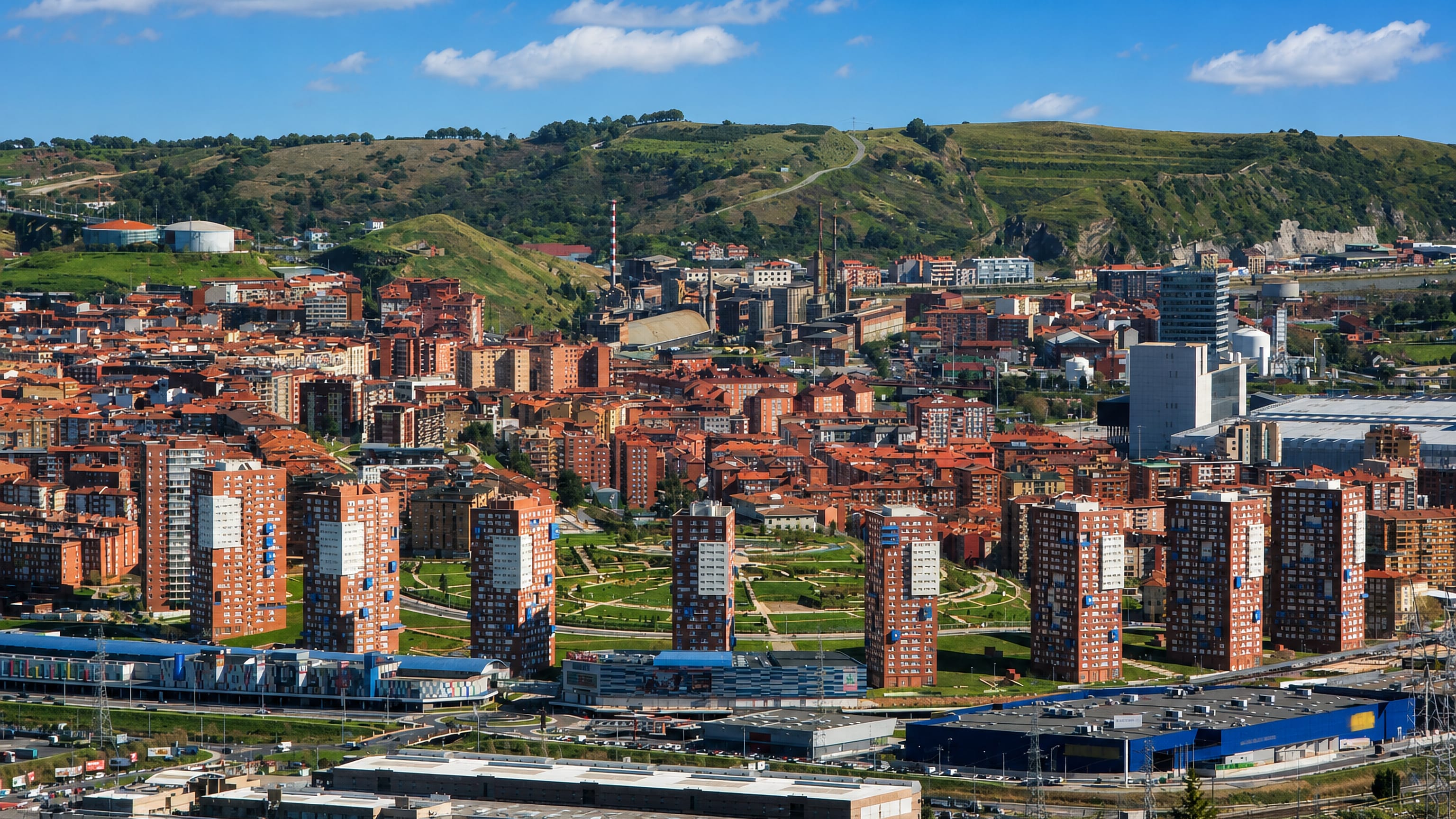 Brick tower blocks, parkland and retail sheds of Barakaldo, Spain, beneath the green hills of the Nervión valley.