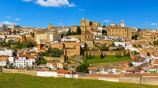 The walled medieval old town of Cáceres with its cathedral tower and Renaissance palaces above green fields.