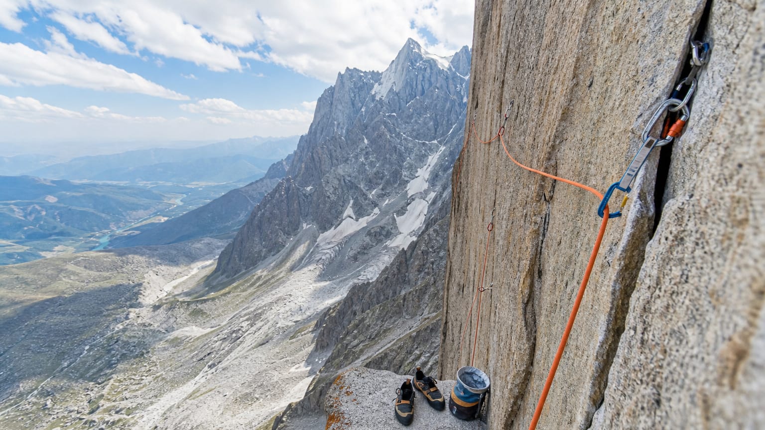 Climbing rope and quickdraws clipped to bolts on a sheer granite wall above a deep alpine valley, snowy peak behind.