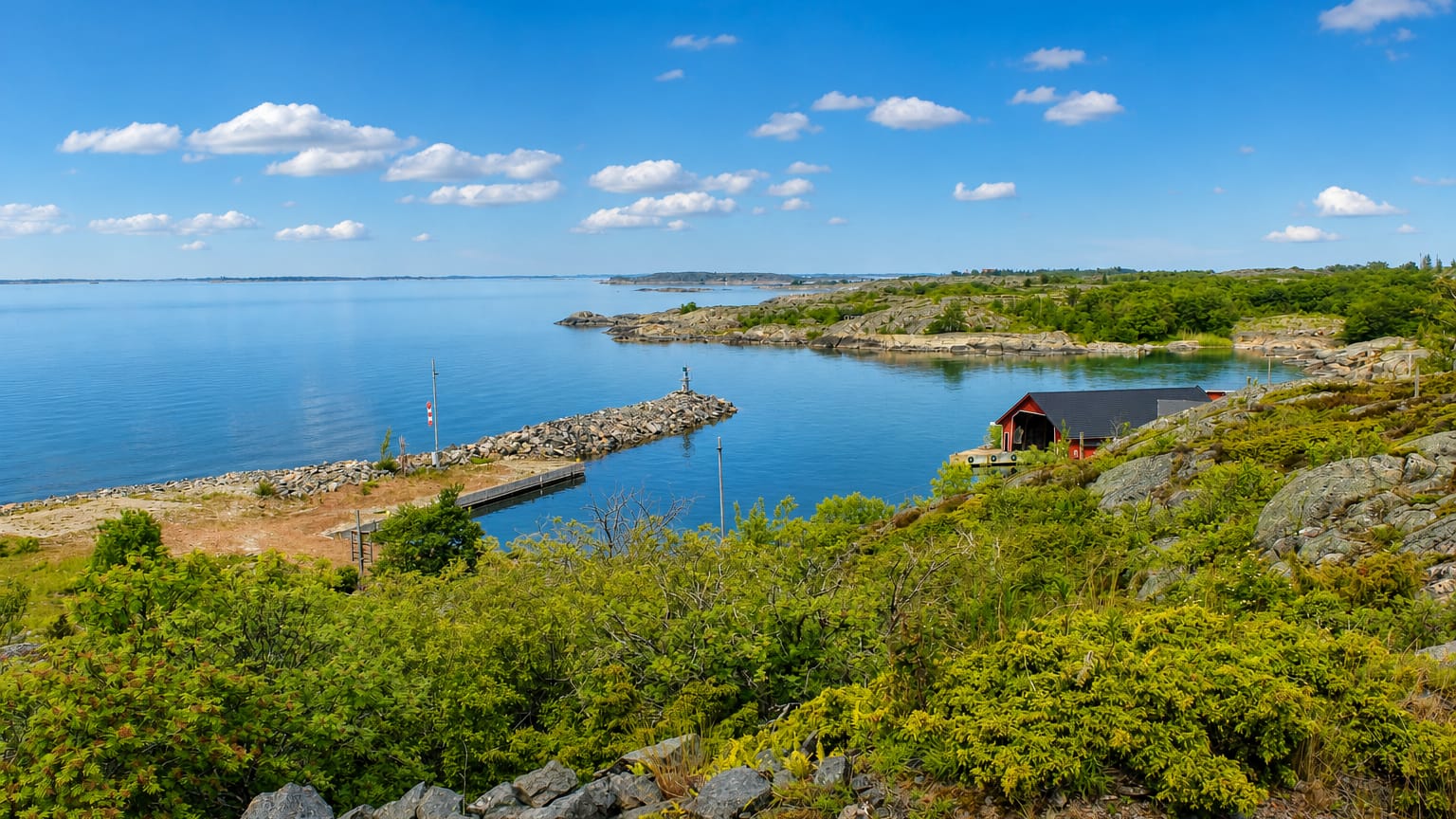 Rocky juniper shoreline and a small harbor with a red boathouse by the blue archipelago sea at Kökar, Finland.