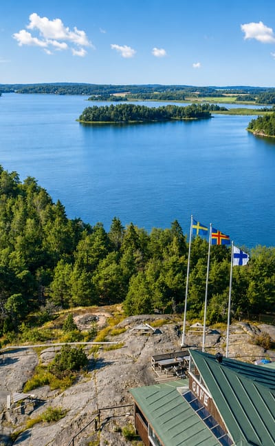 Aerial view over Saltvik's forested Åland archipelago, with Finnish, Swedish and Åland flags on a rocky shoreline outcrop