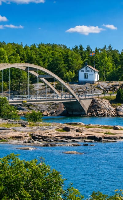 Arched white bridge over blue archipelago water linking forested rocky islets with a flagged cottage in Sund, Åland, Finland