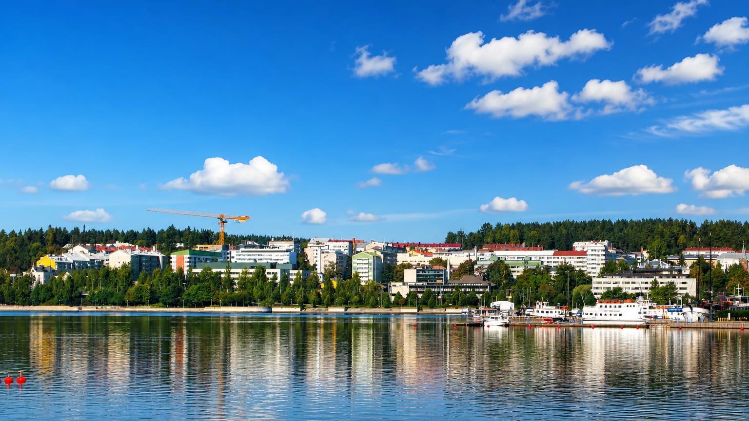 Jyväskylä's low-rise skyline and forested ridges mirrored in the calm waters of Lake Jyväsjärvi under a bright blue sky