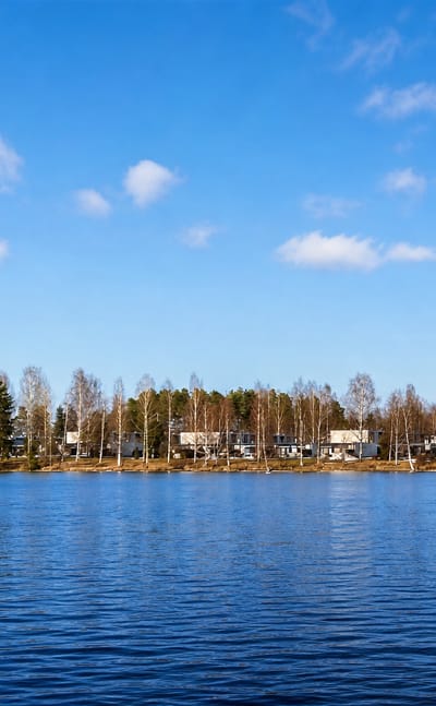 Calm blue lake reflecting the sky, with low residential buildings and birch trees lining the far shore at Lohikoski, Finland.
