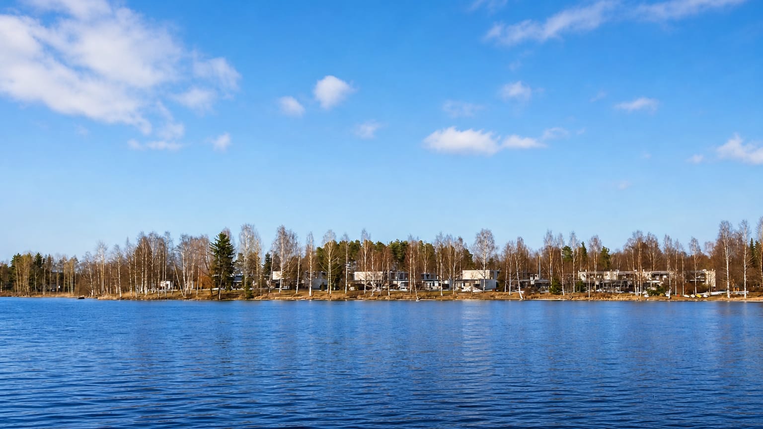 Calm blue lake reflecting the sky, with low residential buildings and birch trees lining the far shore at Lohikoski, Finland.