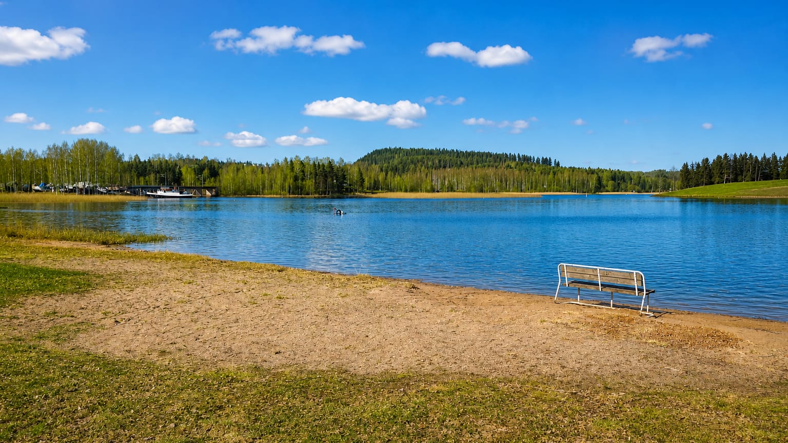 Sandy lakeside beach with a white bench facing a calm blue lake, a small marina and forested shore in Muurame, Finland.