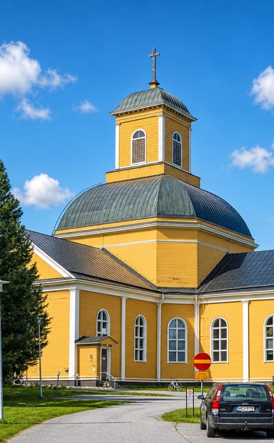 Kuhmo's yellow wooden church with a green dome and cross, framed by tall spruce trees on a sunny day