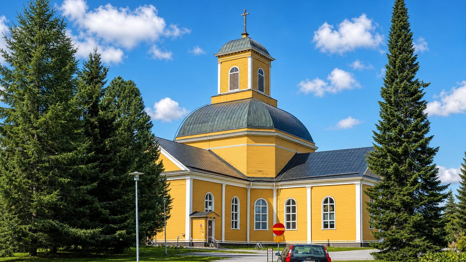 Kuhmo's yellow wooden church with a green dome and cross, framed by tall spruce trees on a sunny day