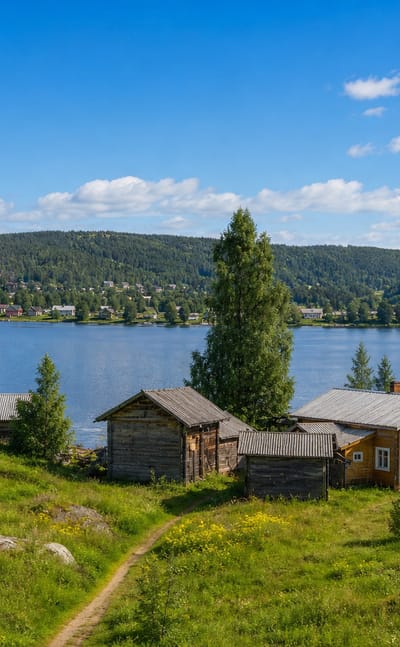 Traditional wooden log buildings on a grassy lakeshore in Paltamo, Finland, with a church spire across the Oulujärvi bay