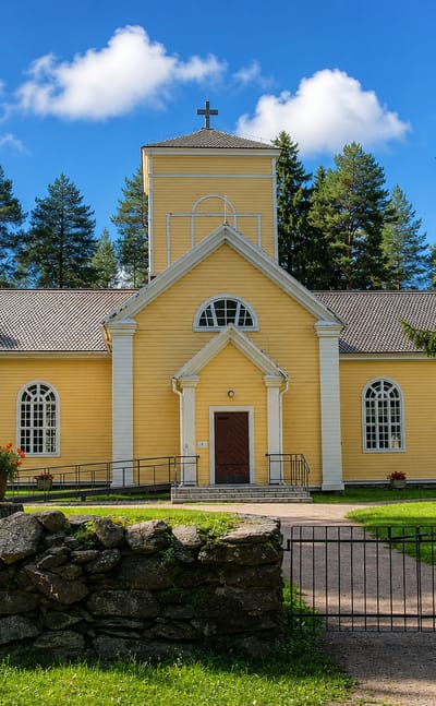 Ristijärvi's yellow wooden church behind a stone wall and iron gate on a summer day.