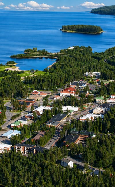 Aerial view of Suomussalmi town centre among boreal forests and blue lakes, with a white water tower and a small boat harbour.