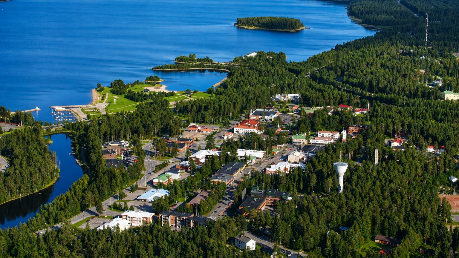 Aerial view of Suomussalmi town centre among boreal forests and blue lakes, with a white water tower and a small boat harbour.
