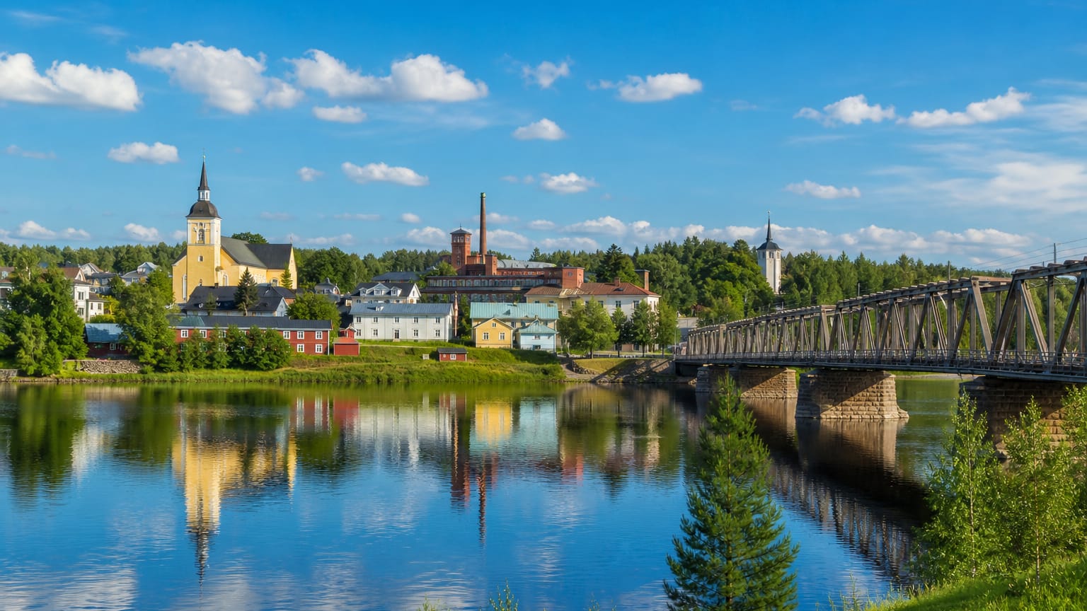 Summer townscape of Vaala, Finland, with a yellow church, brick tower and iron truss bridge mirrored in calm river water