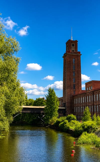 Forssa's red-brick cotton mill and its clock tower beside the calm Loimijoki river, framed by green trees on a sunny day