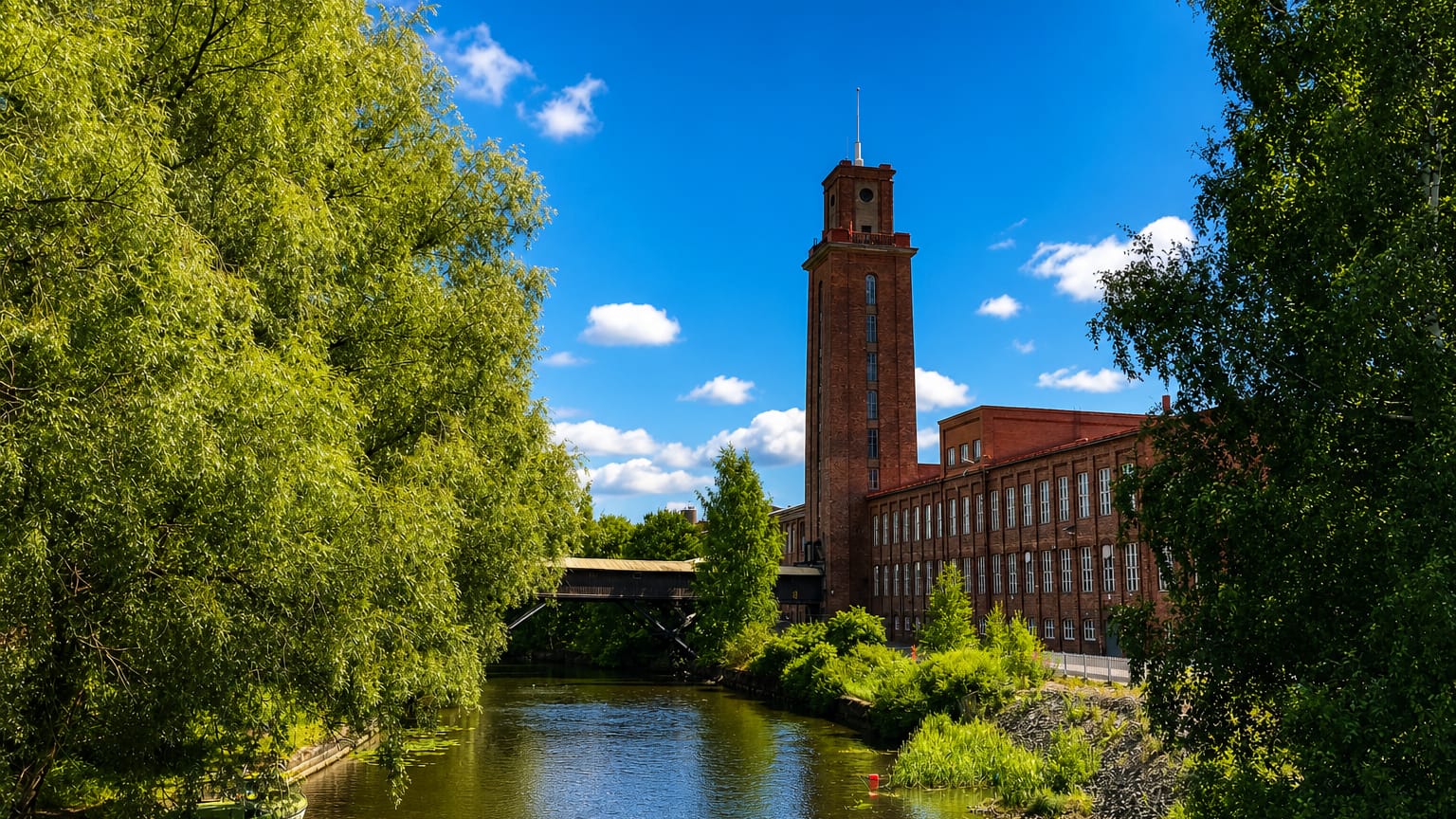 Forssa's red-brick cotton mill and its clock tower beside the calm Loimijoki river, framed by green trees on a sunny day