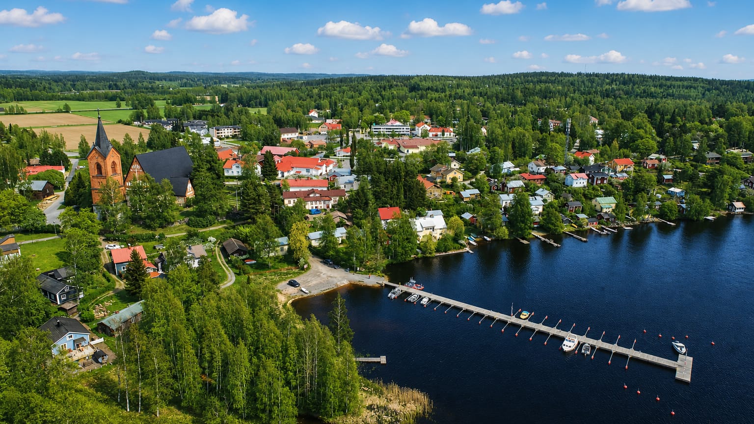 Aerial view of Hauho, Finland: a lakeside village with a church spire, marina and red-roofed houses by forested shores.