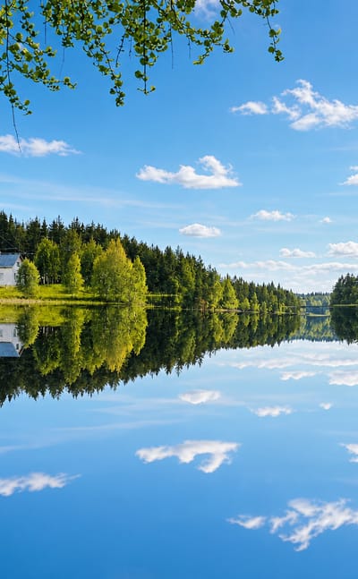 Calm forest-lined lake in Janakkala, Finland, with a pale lakeside cottage mirrored in still blue water