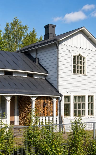 White wooden house with a black metal roof and covered porch on the edge of Oitti, Finland.