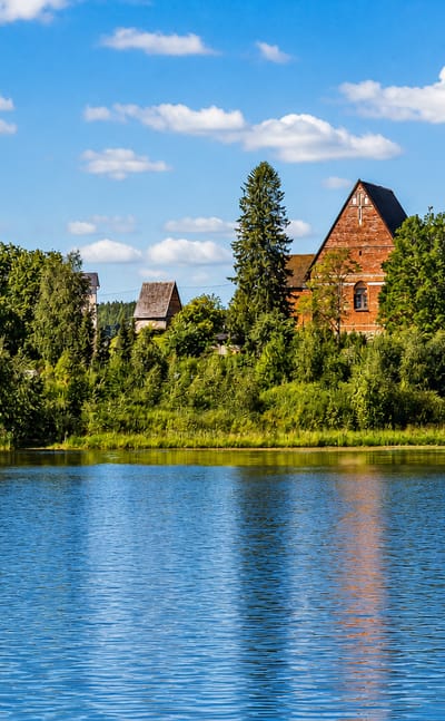 Red-brick church gable rising above a wooded shoreline across a calm lake at Parola, Finland, under a bright summer sky.
