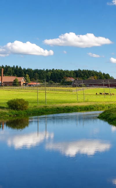 Calm river winding past green pastures and farm buildings in rural Ypäjä, Kanta-Häme, Finland.