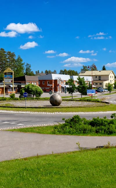 Sunny roundabout with a stone sphere sculpture and low brick and timber buildings in central Perho, Finland.