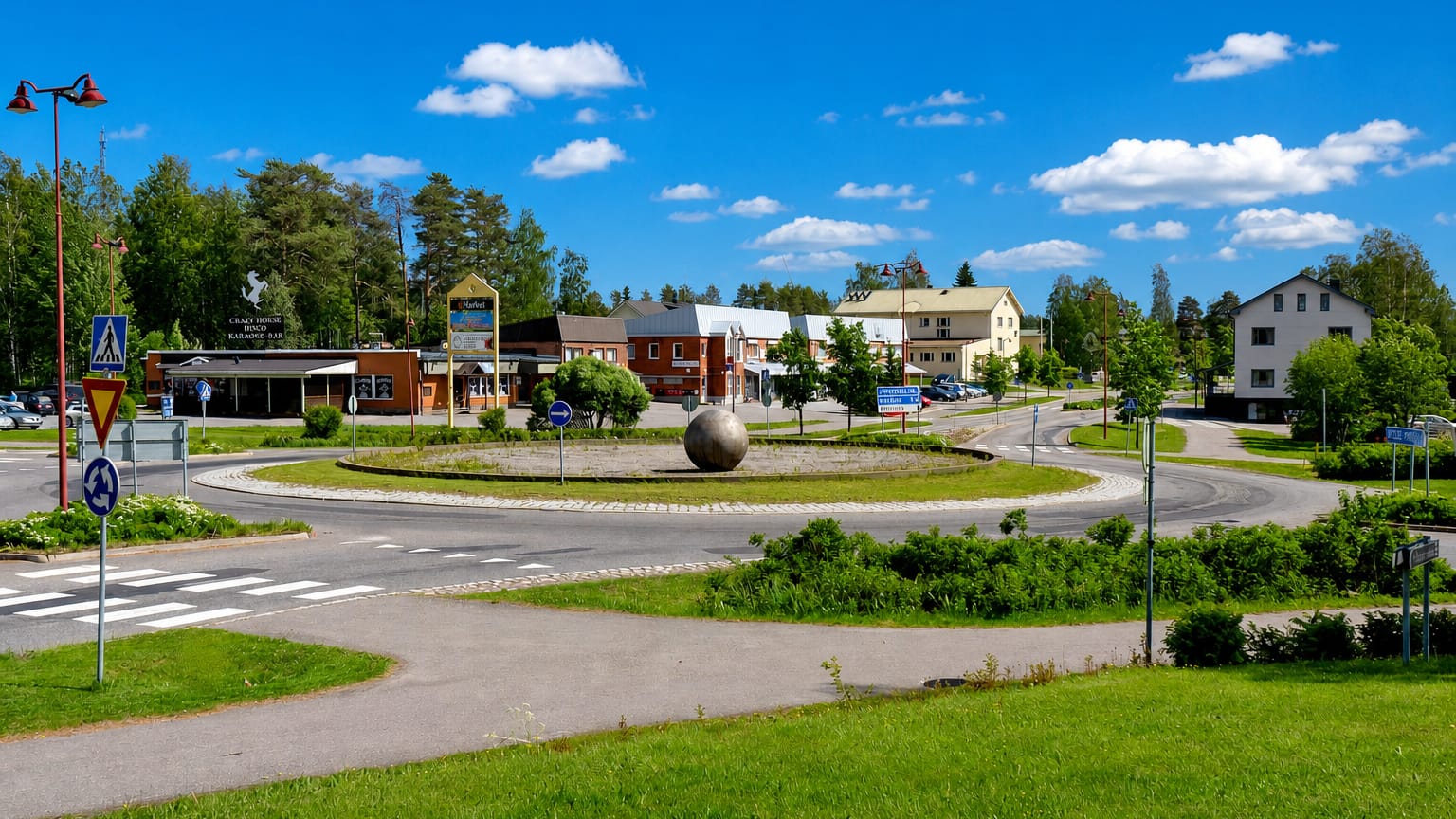 Sunny roundabout with a stone sphere sculpture and low brick and timber buildings in central Perho, Finland.