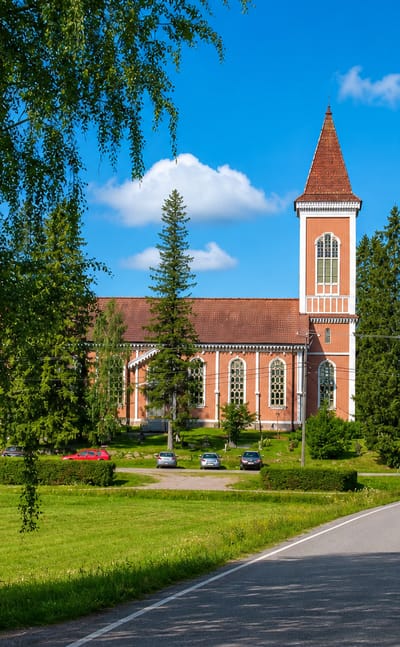 Red-brick church with a tall spire beside a curving road in Toholampi under a bright blue sky.