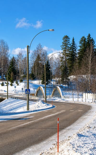 Snow-lined road curving toward a white arched bridge among birch trees at Broby