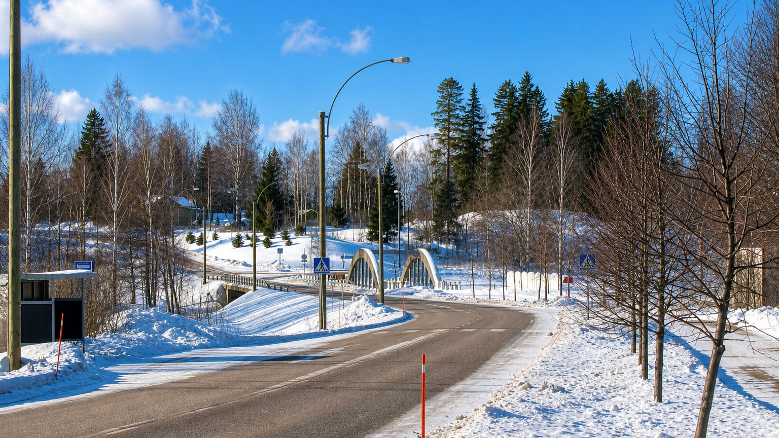 Snow-lined road curving toward a white arched bridge among birch trees at Broby